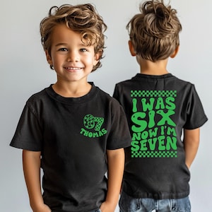 May include: Black t-shirts with green graphics. One shirt has "67 Thomas" on the front. The other shirt has "I WAS SIX NOW I'M SEVEN" on the back. The shirts are for a child's birthday.