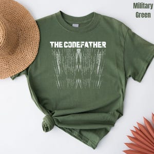May include: Military green t-shirt with the text "THE CODEFATHER" above a white graphic resembling the letter "W" made of vertical lines. The shirt is knotted at the bottom. A straw hat and a decorative leaf are in the background.