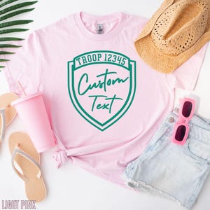 A pink t-shirt with a green shield-shaped logo, a straw hat, sunglasses, and flip flops, all arranged on a white background.