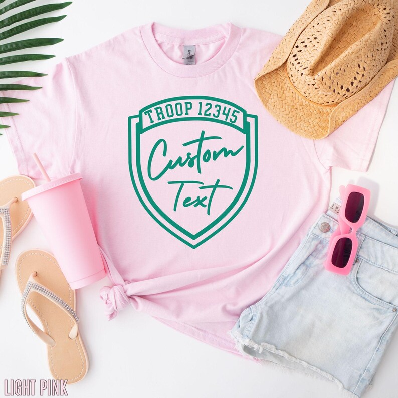A pink t-shirt with a green shield-shaped logo, a straw hat, sunglasses, and flip flops, all arranged on a white background.