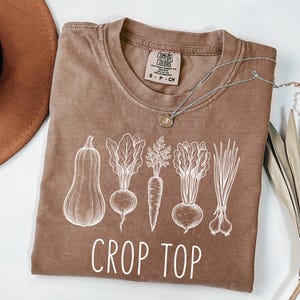 May include: A brown short-sleeve t-shirt featuring a white line-art graphic of a butternut squash, a beet, a carrot, a turnip, and green onions. The text CROP TOP is printed in white below the vegetables. The shirt has a Comfort Colors label.