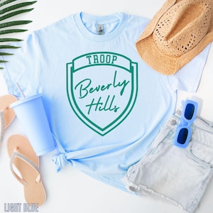 A light blue T-shirt with a green shield-shaped logo, a straw hat, sunglasses, flip flops, and a cup are arranged on a white background.