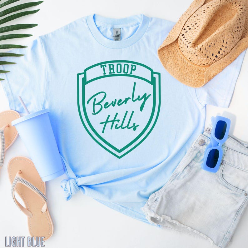 A light blue T-shirt with a green shield-shaped logo, a straw hat, sunglasses, flip flops, and a cup are arranged on a white background.