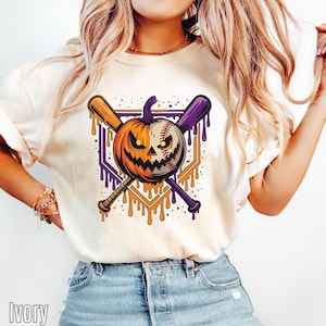 May include: An ivory t-shirt featuring a graphic print of a jack-o'-lantern pumpkin merged with a baseball, positioned in front of two crossed baseball bats. The design includes dripping orange and purple paint effects and a geometric frame.