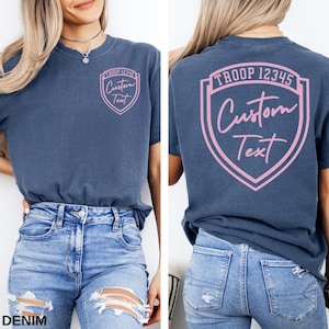 A woman wearing a blue t-shirt with a pink shield design and text on the back, paired with distressed jeans.