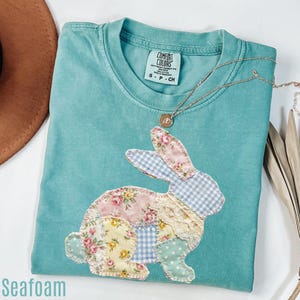 Pode incluir: Camiseta verde-água com um design de coelho de retalhos. O coelho é feito de tecidos florais, xadrez e de bolinhas. Um colar de ouro com um pingente repousa sobre a camisa. A camisa tem a etiqueta da marca Comfort Colors.