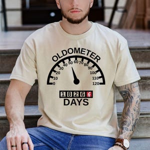 May include: Beige t-shirt with a black "OLDOMETER" graphic. The graphic resembles a speedometer, with the needle pointing to 60. Below the speedometer is the number "18264" and the word "DAYS".