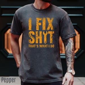 May include: A charcoal gray short-sleeve t-shirt featuring distressed yellow text that reads I FIX SHYT with a wrench replacing the letter Y, followed by the text THAT'S WHAT I DO below it. The shirt has a crew neckline and a standard fit.