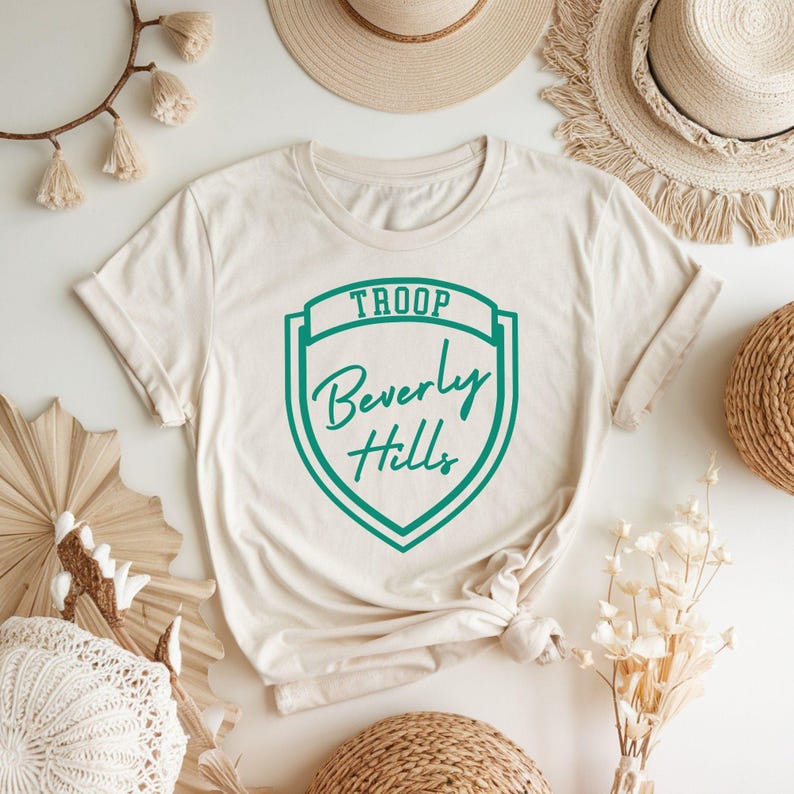 a white T-shirt with a green shield design that says "Troop Beverly Hills" printed on it, along with a straw hat and some decorative elements such as a fan and flowers.