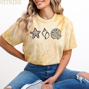 May include: Yellow t-shirt with a tie-dye effect, featuring black line art of a starfish, a conch shell, and a scallop shell. The word "CITRINE" is printed at the top. The model is wearing blue jeans.