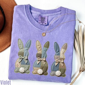 Pode incluir: Uma camiseta Comfort Colors violeta com três coelhos aplicados com desenhos de patchwork. Cada coelho tem um laço e uma cauda de pompom branca. Um colar de ouro com um pingente está drapeado sobre a camisa. A palavra "Violet" está na parte inferior esquerda.