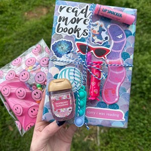 May include: A rectangular gift package wrapped in purple and blue scale-patterned paper, featuring stickers of a cow, sunglasses, and text reading read more books and sorry i was reading. It includes a Lip Smacker, hand sanitizer, and a small fidget toy.