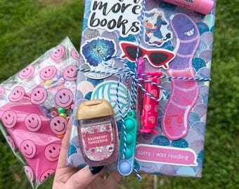 May include: A rectangular gift package wrapped in purple and blue scale-patterned paper, featuring stickers of a cow, sunglasses, and text reading read more books and sorry i was reading. It includes a Lip Smacker, hand sanitizer, and a small fidget toy.