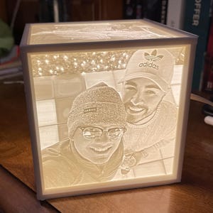May include: A white cube-shaped night light with a 3D engraved image of two people. The people are smiling and wearing casual clothing. The person on the left is wearing a beanie and glasses. The person on the right is wearing a hat with the Adidas logo. The image is illuminated from behind, creating a soft glow.