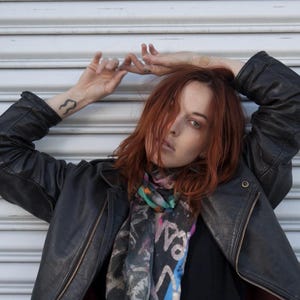 Può includere: Una donna indossa una giacca di pelle nera con una sciarpa a stampa astratta colorata. La sciarpa ha una varietà di colori tra cui rosa, blu, verde e bianco.