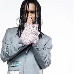 Puede incluir: Un hombre con un blazer gris con parches de tela con estampados coloridos en los bolsillos. Lleva una camisa blanca con estampado floral y pantalones negros. Tiene trenzas negras largas y tiene las manos juntas frente a su cara.