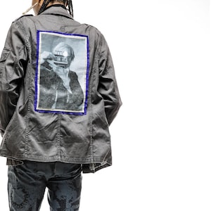 May include: A gray utility jacket with a black and white photo of a person on the back. The photo is framed with a blue border. The person in the photo is wearing a hat and has their hand over their face. The jacket has a zipper closure and two pockets on the front.