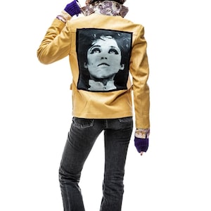 May include: Mustard-colored leather jacket with a black and white portrait on the back. The jacket is worn with dark gray jeans, black boots, a patterned scarf, and purple gloves. The image is set against a white background.