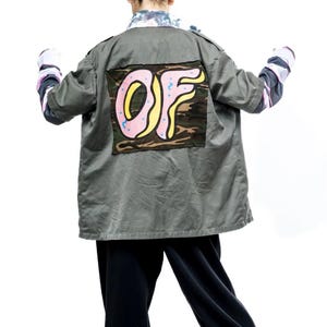 Puede incluir: Una chaqueta verde de utilidad con estampado de camuflaje y un gráfico de donut rosa y amarillo con las letras "OF" en la espalda.