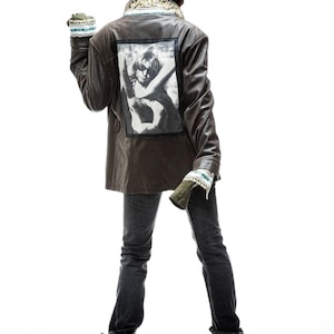 May include: A brown leather jacket with a graphic print on the back. The jacket has a collar and long sleeves. The person is wearing blue jeans and black boots.