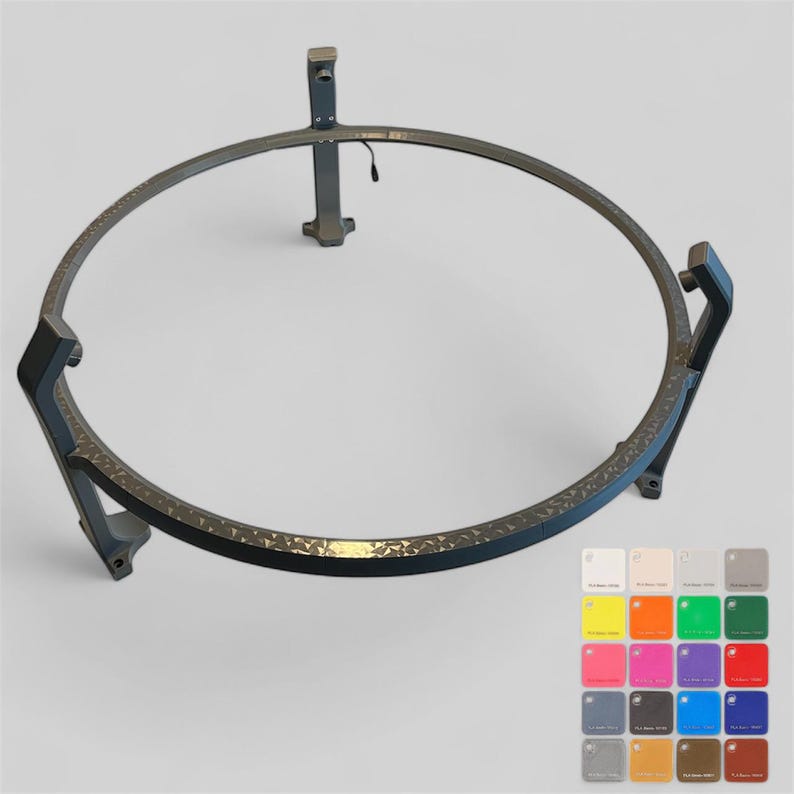Op de afbeelding: Een donkergrijze metalen vuurkorfring met een getextureerd oppervlak. De ring is cirkelvormig met drie poten en een steunpaal. Een kleurenpalet met verschillende tinten bevindt zich rechtsonder.