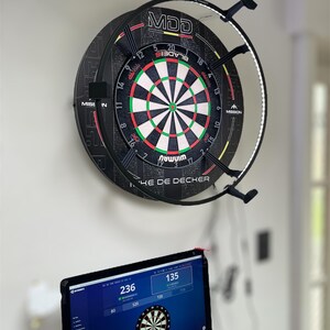 May include: A dartboard with a black and red design, featuring a white, green, and red target, is mounted on a wall. The dartboard has the text "MDD" and "MISSION". A computer monitor below displays a dart game interface with scores and a dartboard graphic.