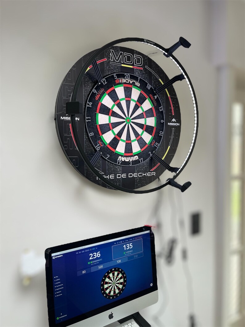 May include: A dartboard with a black and red design, featuring a white, green, and red target, is mounted on a wall. The dartboard has the text "MDD" and "MISSION". A computer monitor below displays a dart game interface with scores and a dartboard graphic.