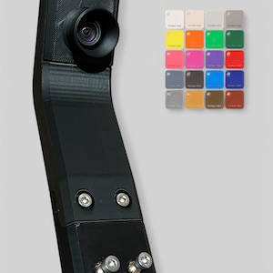 Op de afbeelding: Een zwarte camera met een lens, gemonteerd op een gebogen zwarte arm. De arm is met schroeven bevestigd. Op de achtergrond een kleurenpalet van kleine vierkante kleurstalen, met kleuren als wit, geel, oranje, groen, roze, paars en blauw.
