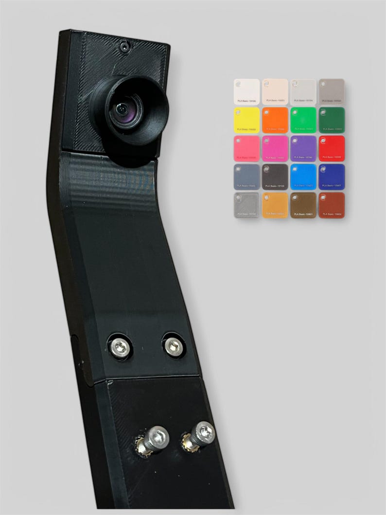 Op de afbeelding: Een zwarte camera met een lens, gemonteerd op een gebogen zwarte arm. De arm is met schroeven bevestigd. Op de achtergrond een kleurenpalet van kleine vierkante kleurstalen, met kleuren als wit, geel, oranje, groen, roze, paars en blauw.