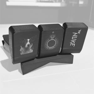 May include: A black desk tidy with three sections. The first section features a dog portrait, the second a crest, and the third reads "THE NUKE". The organiser is made of a dark material and sits on a white surface.