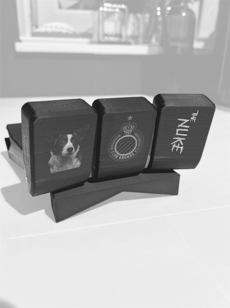 May include: A black desk tidy with three sections. The first section features a dog portrait, the second a crest, and the third reads "THE NUKE". The organiser is made of a dark material and sits on a white surface.