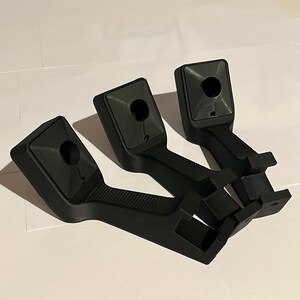 May include: Three black plastic brackets, each with a textured handle and a square top featuring a circular hole. The brackets have a rectangular base with two small, square protrusions. They are on a white surface.