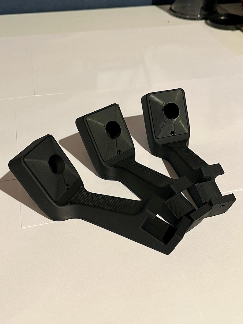 May include: Three black plastic brackets, each with a textured handle and a square top featuring a circular hole. The brackets have a rectangular base with two small, square protrusions. They are on a white surface.