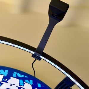 May include: A black, curved LED light with a rectangular head and a ribbed handle is attached to a blue and white object. The light is emitting a bright white glow. The object has the word "FAN" in large white letters.
