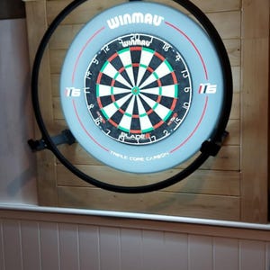 Op de afbeelding: Een Winmau dartbord met een zwarte metalen rand, gemonteerd op een houten muur. Het dartbord is lichtblauw met een zwart, wit en groen doelgebied. De tekst "WINMAU" is zichtbaar bovenaan.