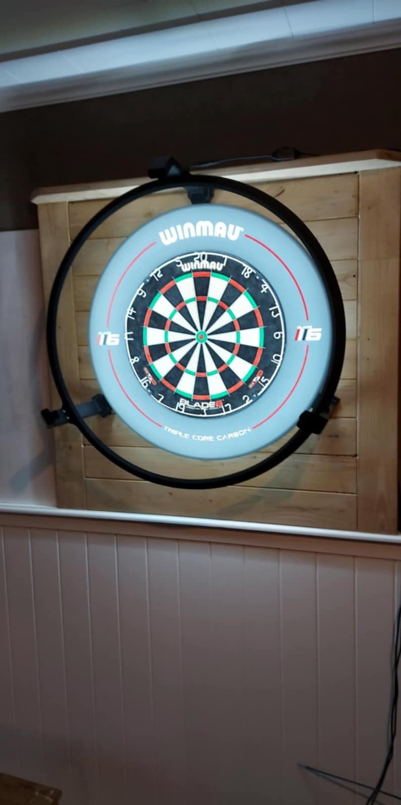 Op de afbeelding: Een Winmau dartbord met een zwarte metalen rand, gemonteerd op een houten muur. Het dartbord is lichtblauw met een zwart, wit en groen doelgebied. De tekst "WINMAU" is zichtbaar bovenaan.