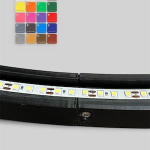 Op de afbeelding: Een zwart, gebogen LED-armatuur met een witte LED-strip. Een kleurenpalet met kleine vierkante kleurstalen bevindt zich in de linkerbovenhoek. De kleurstalen omvatten wit, beige, geel, oranje, groen, roze, paars, rood, grijs, bruin, blauw en donkerrood.