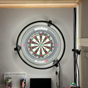 Op de afbeelding: Een Winmau Blade 5 dartbord met een zwarte rand is aan een grijze muur bevestigd. Het dartbord heeft een klassiek ontwerp met rode, groene en witte segmenten. De afbeelding toont ook een lamp en andere woondecoratie.