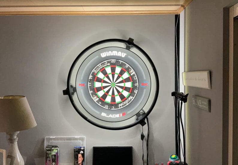 Op de afbeelding: Een Winmau Blade 5 dartbord met een zwarte rand is aan een grijze muur bevestigd. Het dartbord heeft een klassiek ontwerp met rode, groene en witte segmenten. De afbeelding toont ook een lamp en andere woondecoratie.