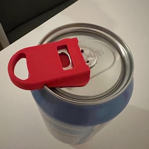 Puede incluir: Un abrelatas rojo está colocado en la parte superior de una lata de bebida azul y plateada. El abrelatas tiene un asa y una abertura curva. La lata está parcialmente abierta, lista para beber.