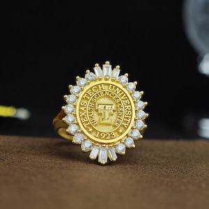 May include: A gold ring with a circular design featuring the Texas Tech University Health Sciences Center logo and the year 1923. The ring is adorned with a halo of clear cubic zirconia stones.