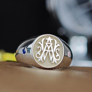 May include: A silver signet ring with a light beige circular face featuring an ornate design with the letters "A" and "W" and a crown. The ring has a blue gemstone on one side and a pink gemstone on the other.