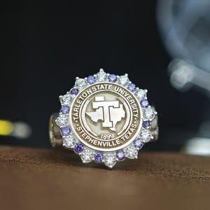 May include: A silver ring with a purple and white stone setting. The ring features the Tarleton State University logo, which includes the words "Tarleton State University", "Stephenville, Texas", and "1899".