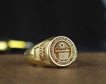 May include: A gold-toned signet ring featuring a circular face with an engraved sun, an open book, and the text "TERRAS IRRADIENT" surrounded by "SIGILL COLL AMHERST MASS NOV ANG MDCCCXXV". The side of the ring is engraved with the year "2025".
