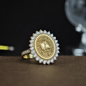 May include: A gold ring with the Houston Christian University seal, encircled by clear gemstones. The ring's band also has smaller gemstones. The university seal features a book, cross, and the text "Houston Christian University Texas - 1960."