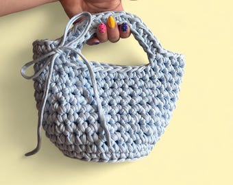 Könnte beinhalten: Hellblaue Häkeltasche mit runder Form und zwei Henkeln. Die Tasche hat ein strukturiertes Stichmuster und einen Kordelverschluss. Die Griffe sind geflochten, und die Tasche wird von einer Hand mit farbenfrohem Nagellack gehalten.