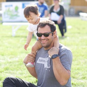 May include: A man wearing sunglasses and a gray t-shirt with the word "home" and a map graphic, carrying a child on his shoulders. The child is wearing a white shirt and looking down. The scene takes place outdoors on a sunny day.