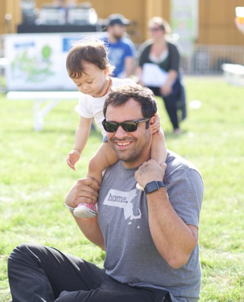 May include: A man wearing sunglasses and a gray t-shirt with the word "home" and a map graphic, carrying a child on his shoulders. The child is wearing a white shirt and looking down. The scene takes place outdoors on a sunny day.