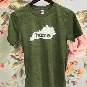 May include: Green t-shirt with a white outline of the state of Kentucky and the word "home" printed in white. The shirt is hanging on a wooden hanger against a floral background.
