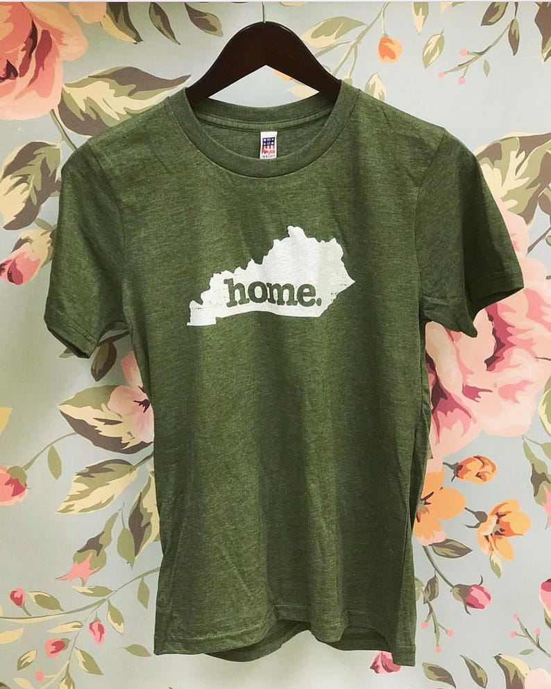 May include: Green t-shirt with a white outline of the state of Kentucky and the word "home" printed in white. The shirt is hanging on a wooden hanger against a floral background.